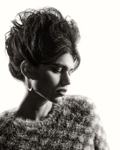 Monochrome portrait of a person with dark hair in an updo wearing hoop earrings and a textured fur-like top against a white backdrop.